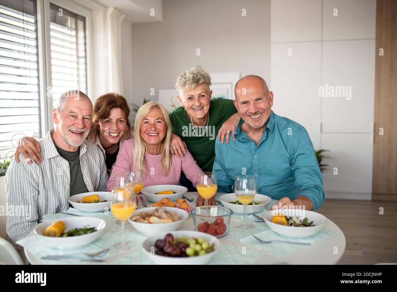 Gruppe älterer Freunde, die in einem Gebäude feiern und beim Essen auf die Kamera schauen. Stockfoto