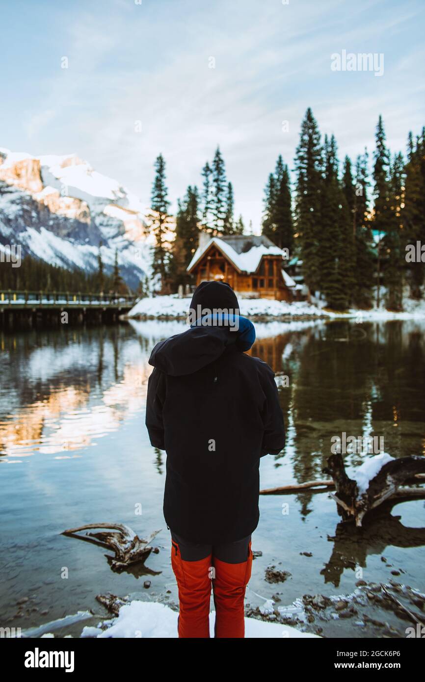 Rückansicht eines nicht erkennbaren männlichen Touristen in Oberbekleidung, der im Winter auf der verschneiten Küste des Emerald Lake Holzhütte und Bergrücken bewundert Stockfoto