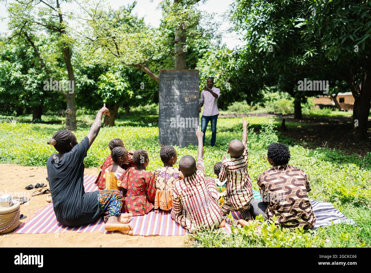 Gemischte Gruppe von schwarzen Kindern, die im Freien sitzen und eifrig freiwillig die Fragen ihres Lehrers während des Unterrichts in einer afrikanischen Gemeinschaftsschule beantworten Stockfoto