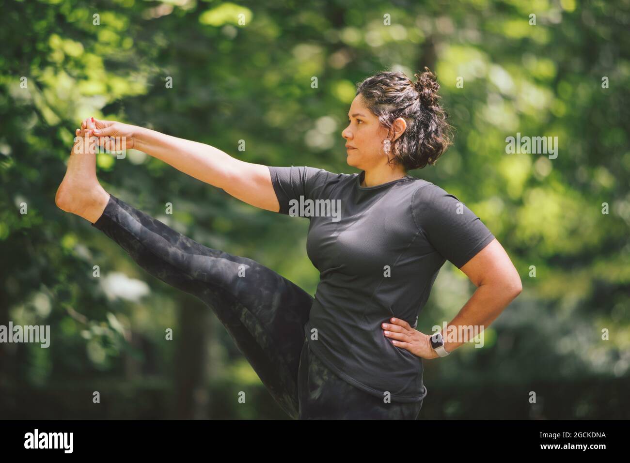 Eine dicke Hündin mit der Hand an der Taille, die in Utthita Hasta Padanguschthasana steht, während sie Yoga praktiziert und im Park nach vorne blickt Stockfoto
