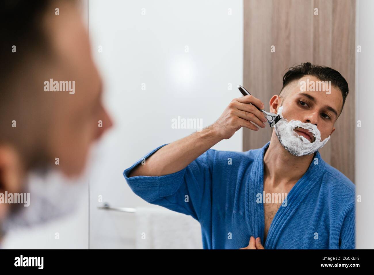 Crop konzentrierte junge ethnische Kerl in blauen Bademantel Blick in  Spiegel und Rasieren mit Rasierer während der täglichen Beauty-Routine im  Bad Stockfotografie - Alamy