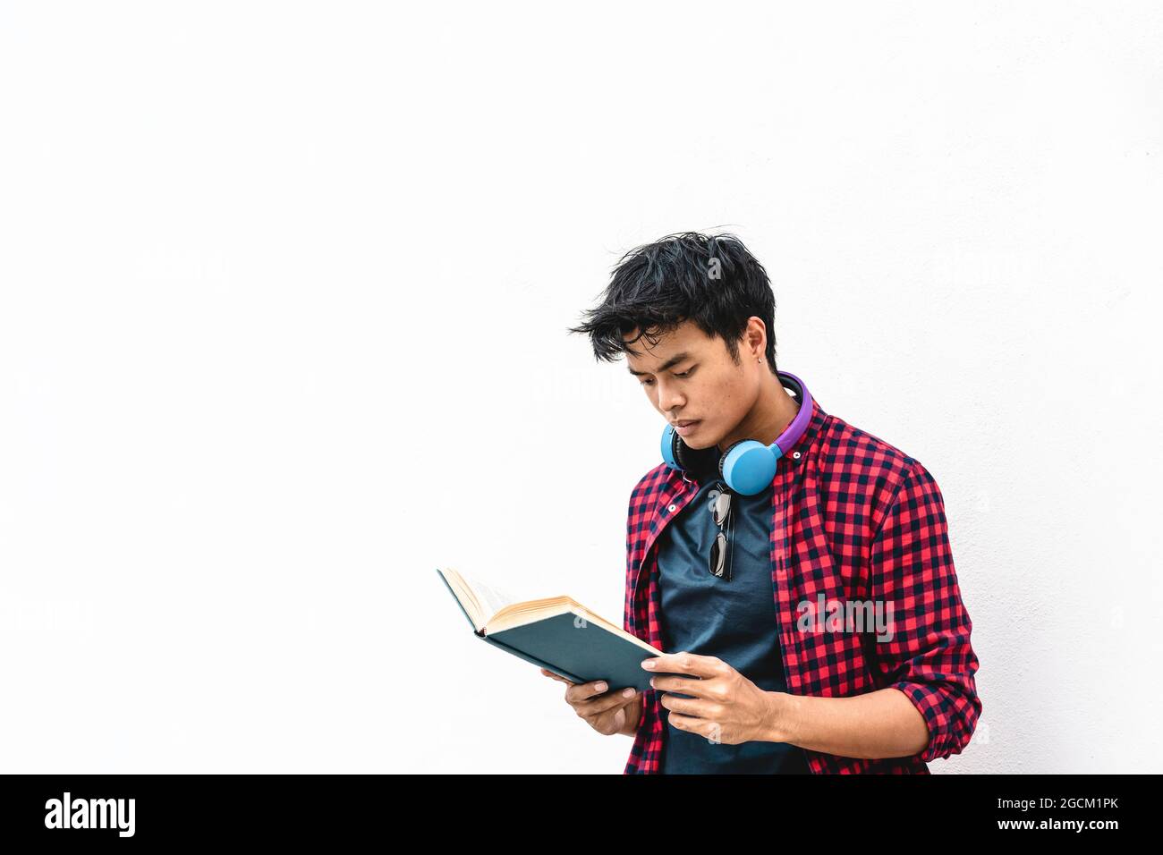 Asiatischer Hipster junger Mann, der ein Buch gegen eine weiße Wand liest - asiatischer Student, der ein Buch in den Händen hält und dies während der Freizeit im Freien liest - Le Stockfoto