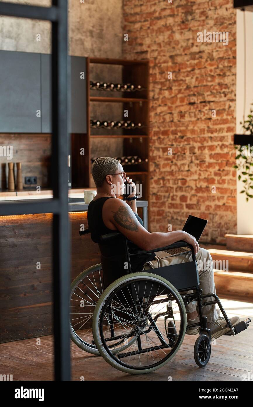 Vertikale Seitenansicht Porträt einer zeitgenössischen tätowierten Frau mit Behinderung mit Laptop während der Arbeit von zu Hause aus Stockfoto