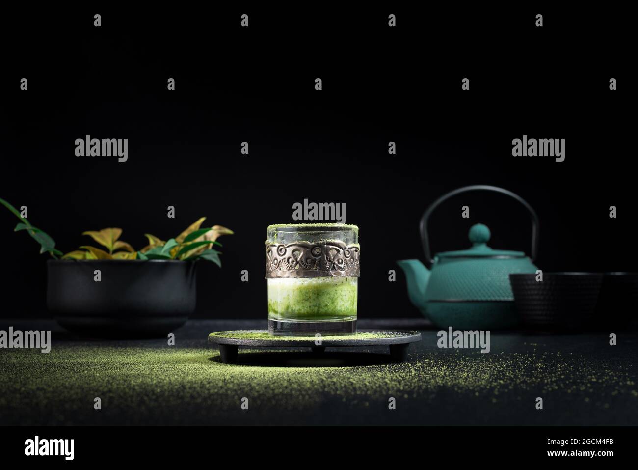 Gesunder grüner Matcha-Tee aus Kräutern, serviert in einer Glastasse mit Metalldekor auf einer Untertasse, die mit Pulver auf dem schwarzen Tisch bestreut ist Stockfoto