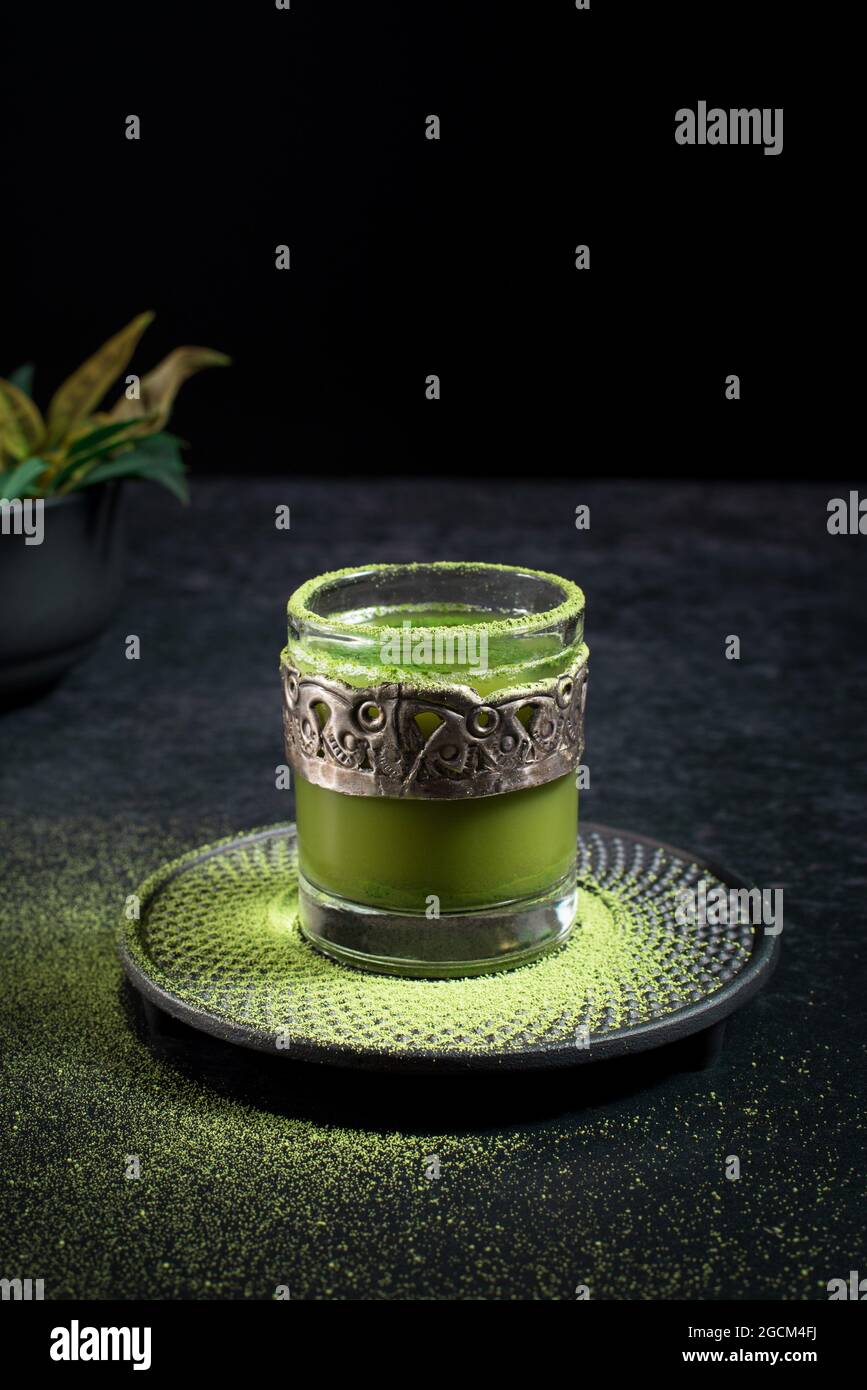 Von oben von gesunden pflanzlichen grünen Matcha-Tee in Glas-Tasse mit Metall-Dekoration auf Untertasse mit Pulver auf schwarzem Tisch bestreut serviert Stockfoto