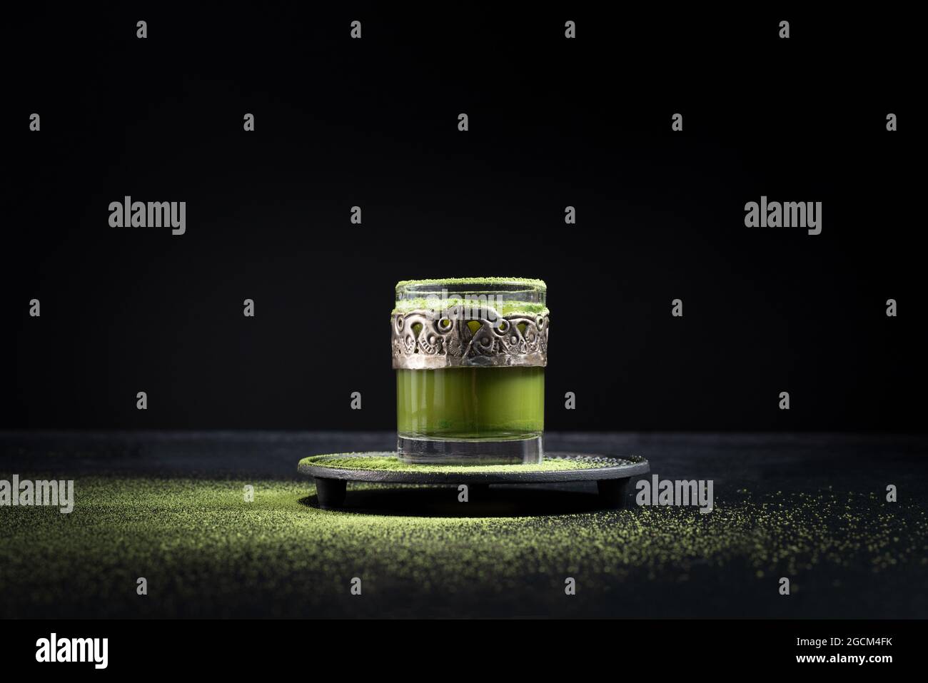 Gesunder grüner Matcha-Tee aus Kräutern, serviert in einer Glastasse mit Metalldekor auf einer Untertasse, die mit Pulver auf dem schwarzen Tisch bestreut ist Stockfoto