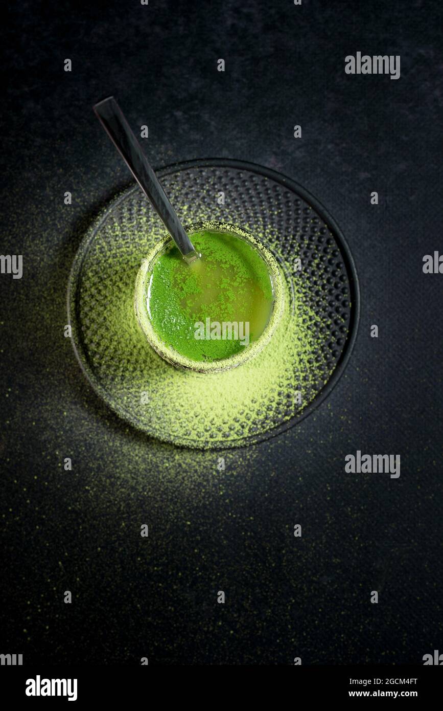 Von oben von gesunden pflanzlichen grünen Matcha-Tee in Glas-Tasse mit Metall-Dekoration auf Untertasse mit Pulver auf schwarzem Tisch bestreut serviert Stockfoto