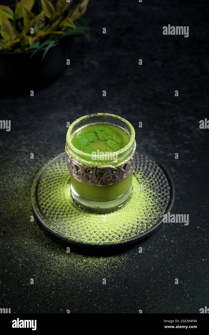 Von oben von gesunden pflanzlichen grünen Matcha-Tee in Glas-Tasse mit Metall-Dekoration auf Untertasse mit Pulver auf schwarzem Tisch bestreut serviert Stockfoto