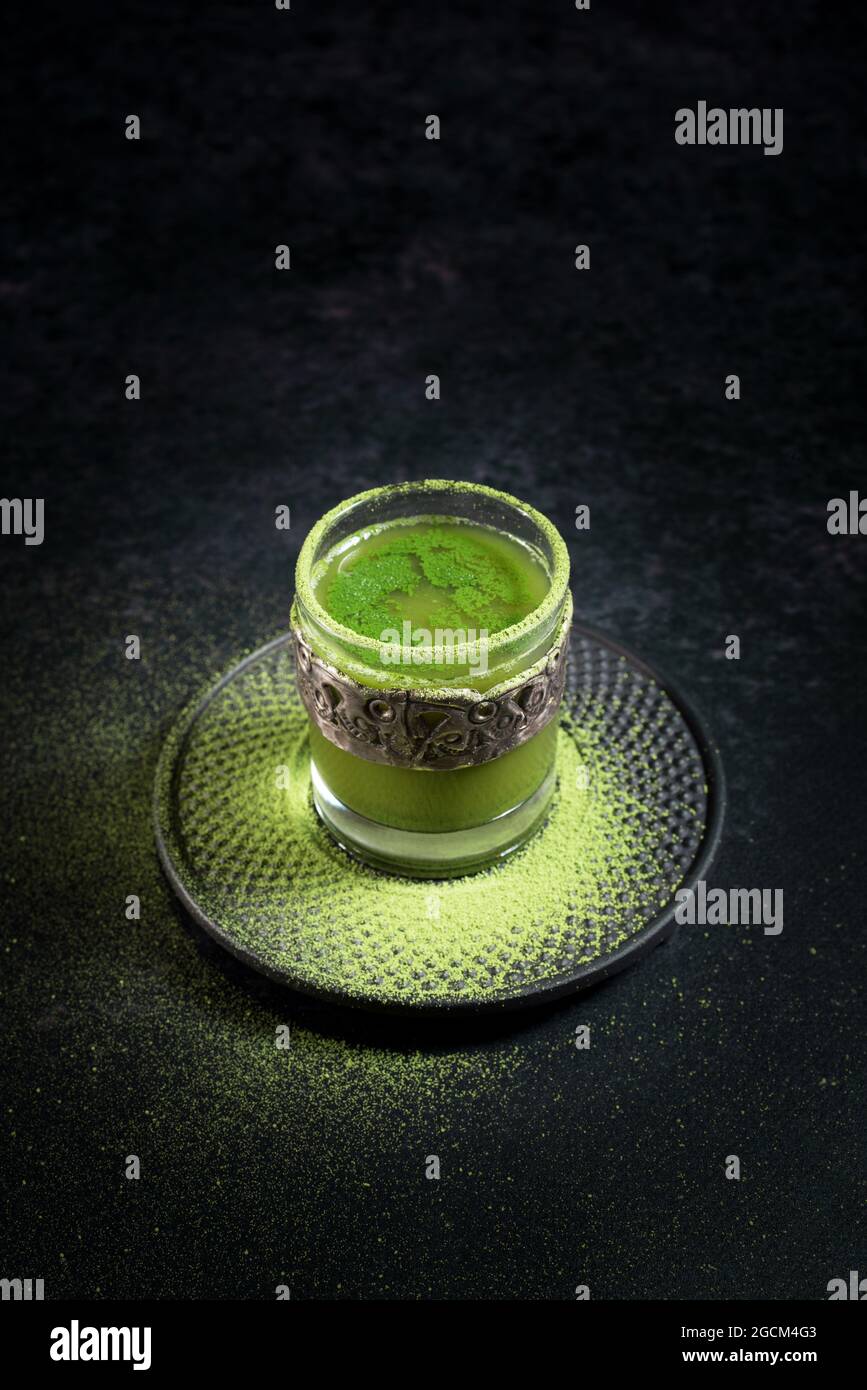 Von oben von gesunden pflanzlichen grünen Matcha-Tee in Glas-Tasse mit Metall-Dekoration auf Untertasse mit Pulver auf schwarzem Tisch bestreut serviert Stockfoto