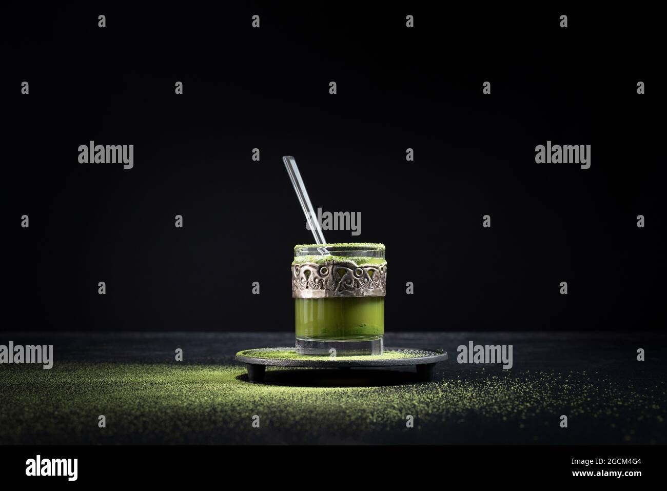 Gesunder grüner Matcha-Tee aus Kräutern, serviert in einer Glastasse mit Metalldekor auf einer Untertasse, die mit Pulver auf dem schwarzen Tisch bestreut ist Stockfoto