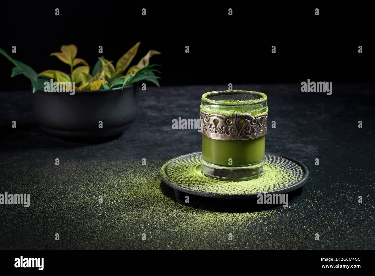 Von oben von gesunden pflanzlichen grünen Matcha-Tee in Glas-Tasse mit Metall-Dekoration auf Untertasse mit Pulver auf schwarzem Tisch bestreut serviert Stockfoto