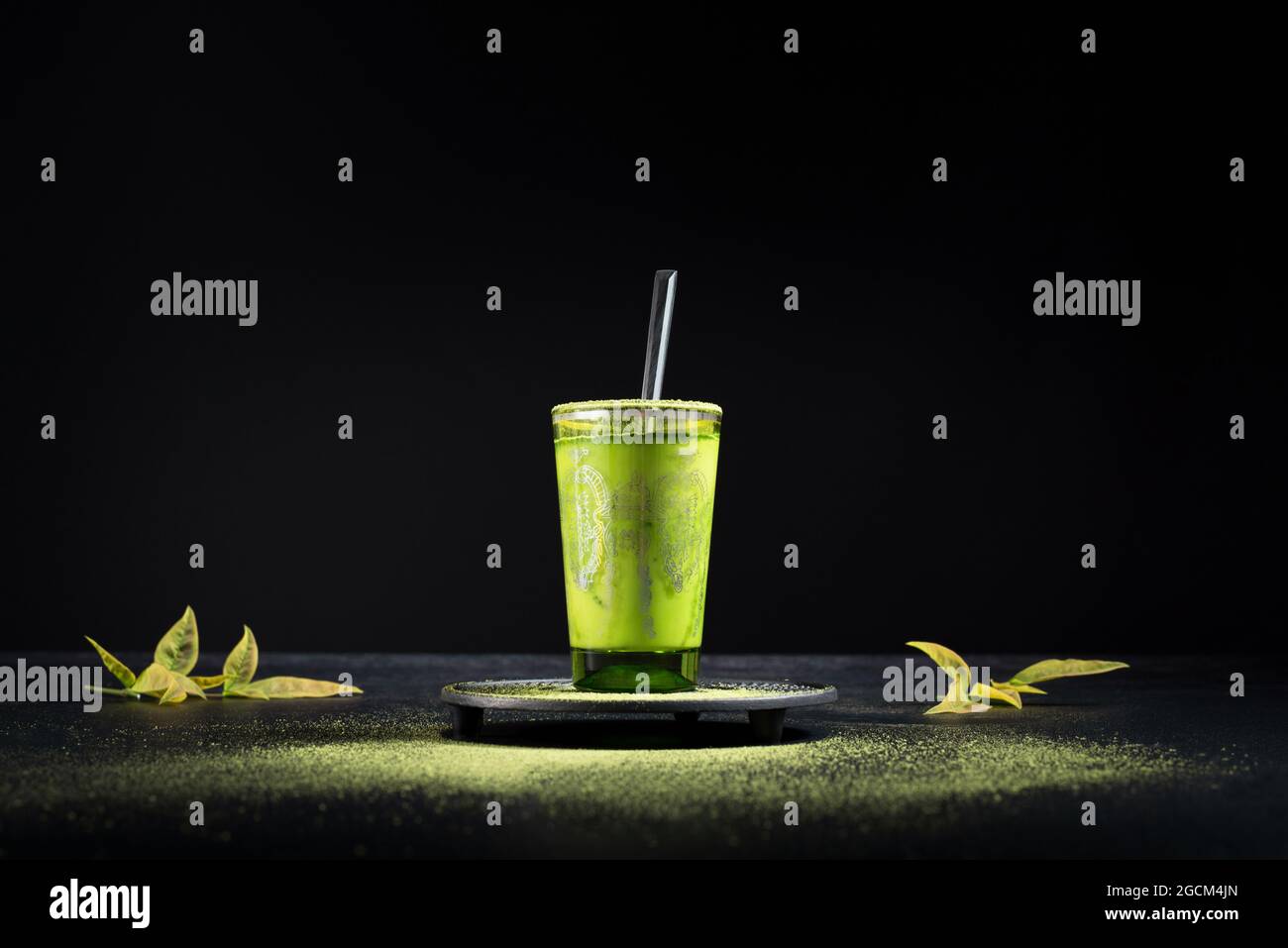 Gesunder grüner Matcha-Tee aus Kräutern, serviert in einer Glastasse mit Metalldekor auf einer Untertasse, die mit Pulver auf dem schwarzen Tisch bestreut ist Stockfoto