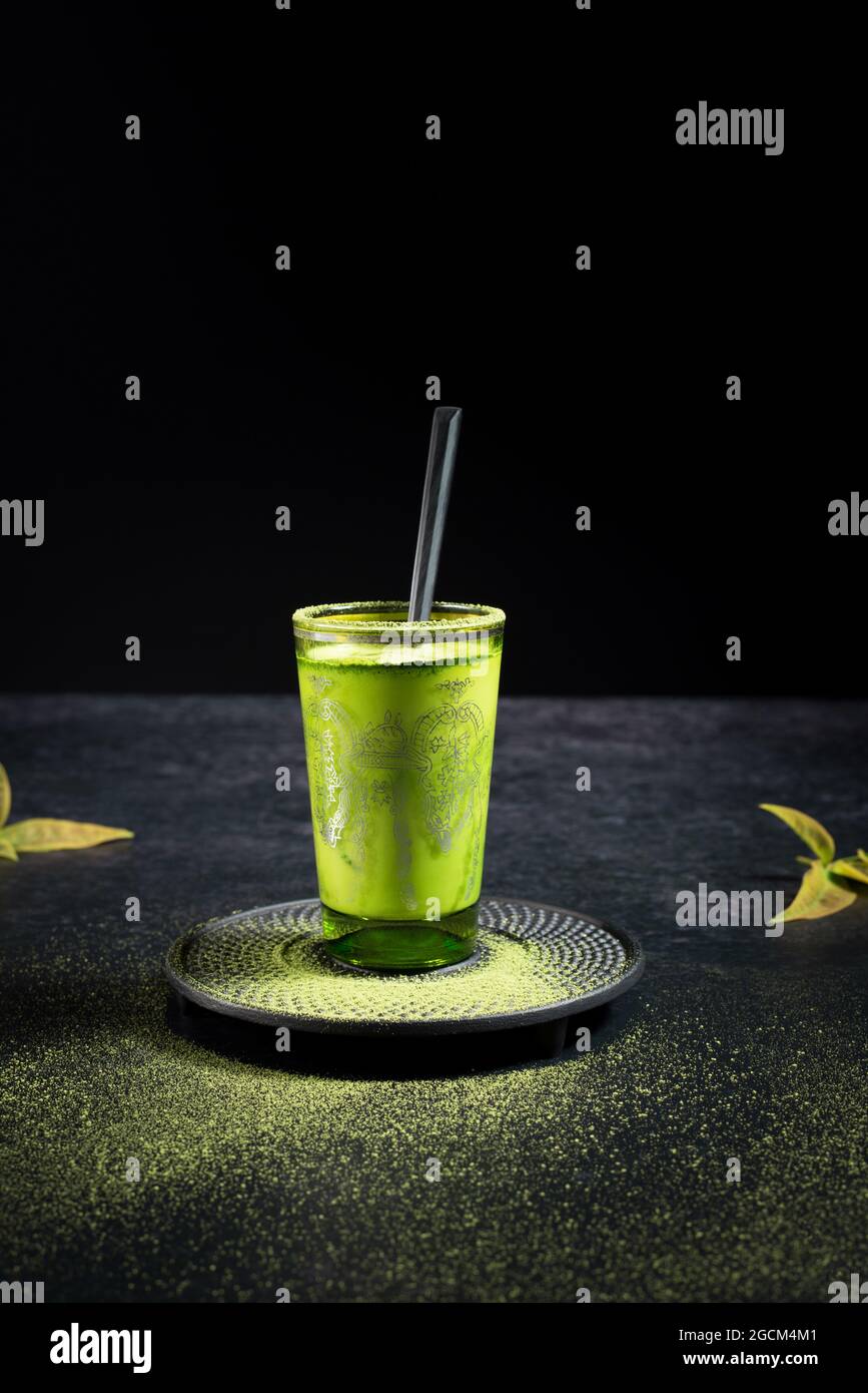Gesunder grüner Matcha-Tee aus Kräutern, serviert in einer Glastasse mit Metalldekor auf einer Untertasse, die mit Pulver auf dem schwarzen Tisch bestreut ist Stockfoto