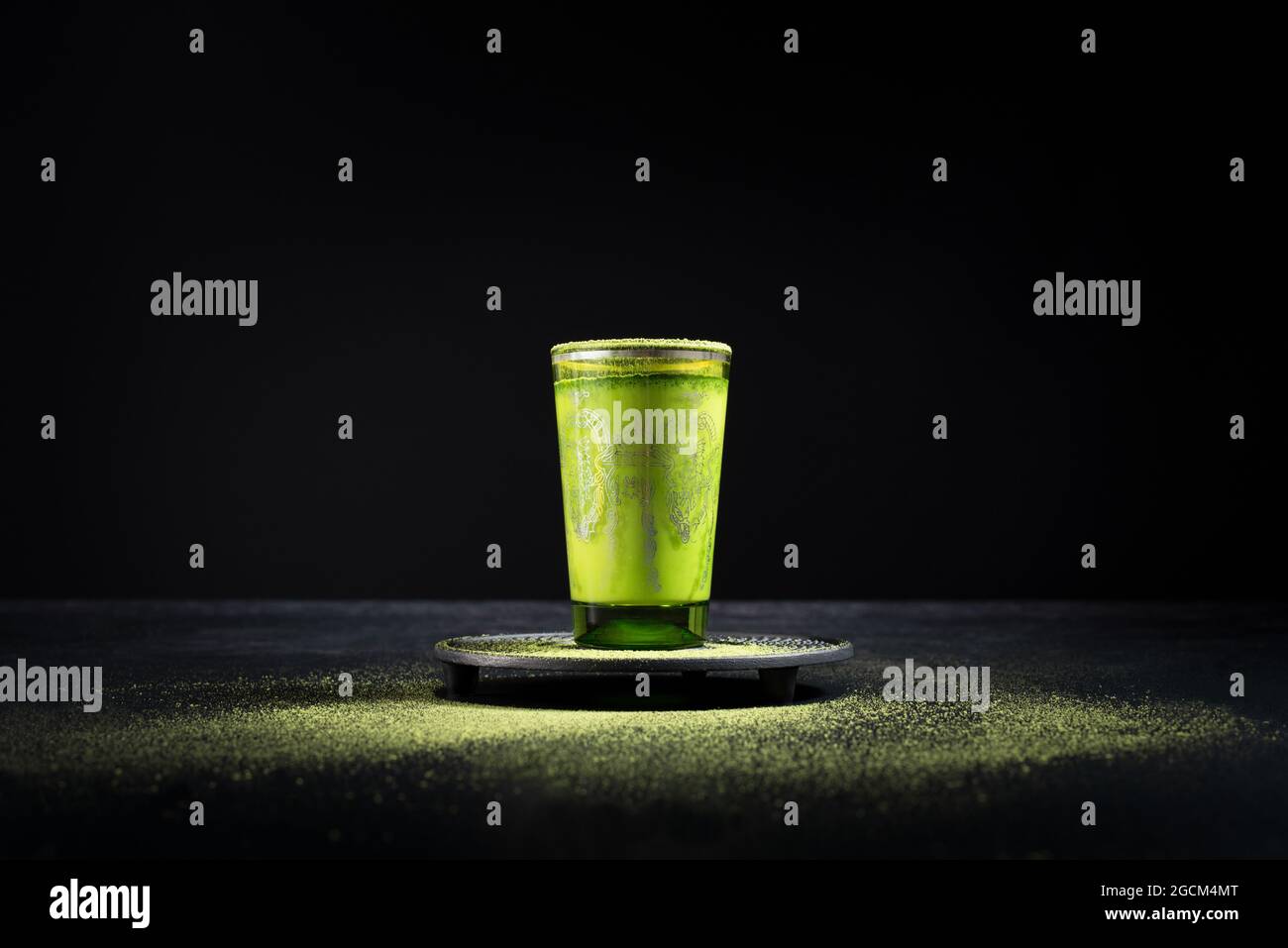 Gesunder grüner Matcha-Tee aus Kräutern, serviert in einer Glastasse mit Metalldekor auf einer Untertasse, die mit Pulver auf dem schwarzen Tisch bestreut ist Stockfoto