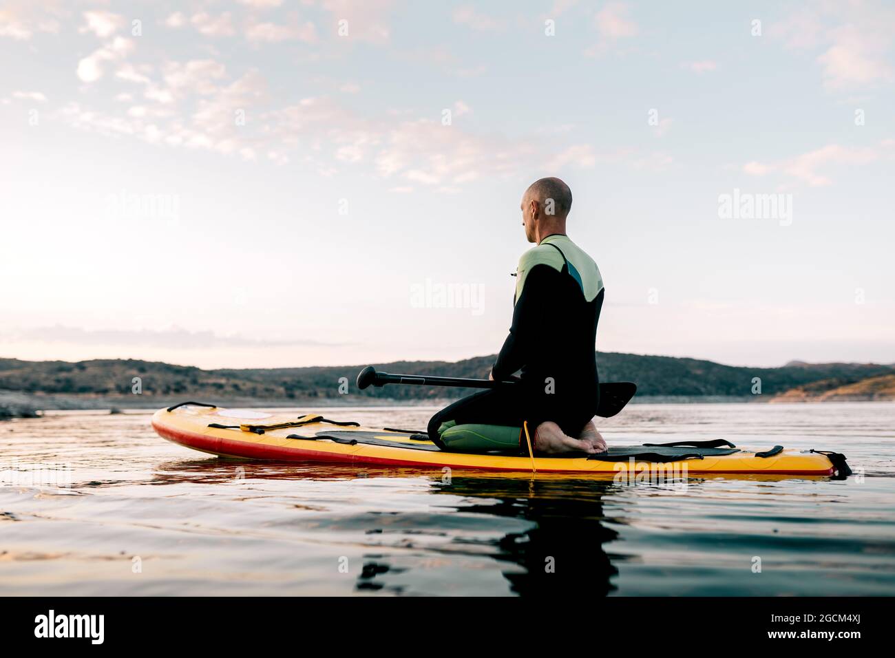 Seitenansicht des ruhigen männlichen Surfers, der in Thunderbolt-Pose auf dem Paddleboard sitzt und am Abend beim Yoga im Meer mediiert Stockfoto