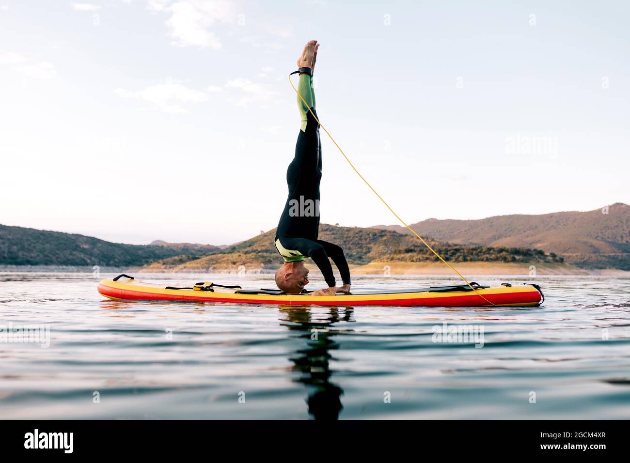 Seitenansicht des männlichen Surfers, der bei Sonnenuntergang Yoga in Kopfstand-Haltung auf dem SUP-Board im Meer praktiziert Stockfoto