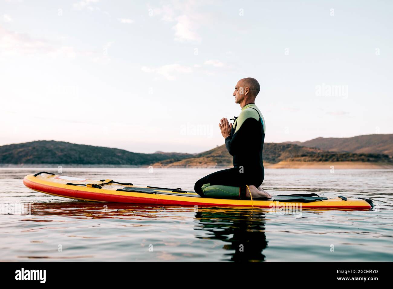 Seitenansicht eines ruhigen männlichen Surfers, der in Thunderbolt-Pose mit Namaste-Händen auf dem Paddleboard sitzt und beim Yoga-Üben im Meer am Abend vermittelt Stockfoto