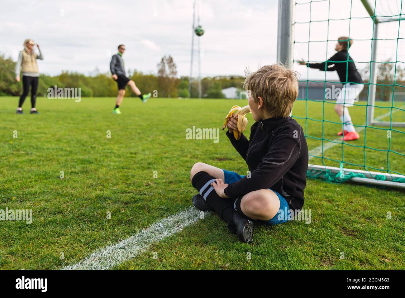 Nicht erkennbarer Teenager, der auf dem Fußballfeld sitzt und Banane isst, während er die Familie beim Fußballspielen anschaut Stockfoto