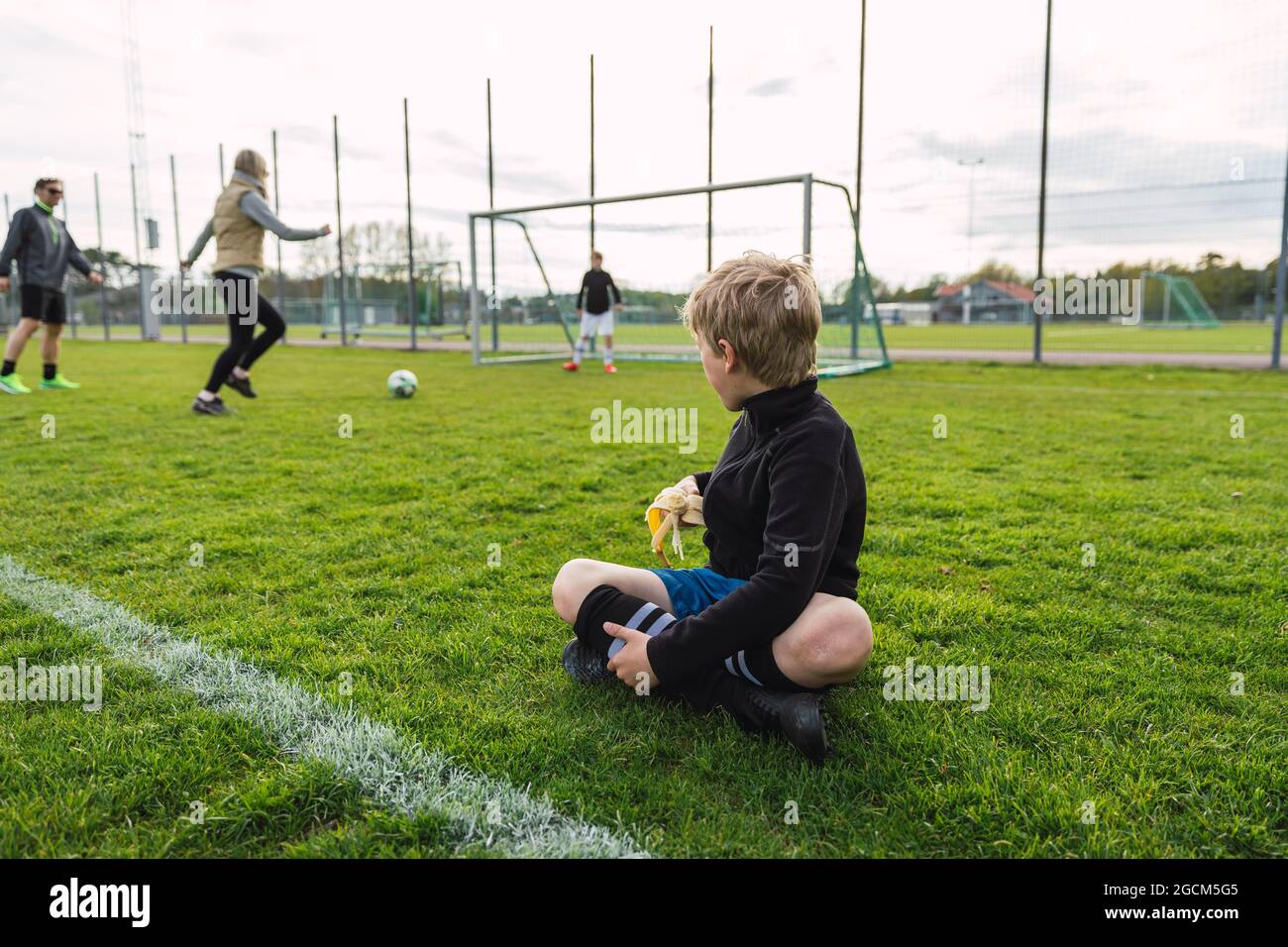 Nicht erkennbarer Teenager, der auf dem Fußballfeld sitzt und Banane isst, während er die Familie beim Fußballspielen anschaut Stockfoto