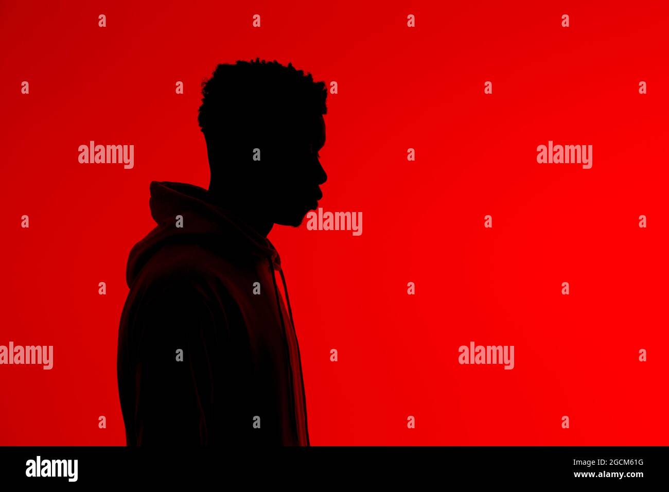 Seitenansicht der Silhouette eines nicht erkennbaren afroamerikanischen Mannes im Hoodie, der auf rotem Hintergrund im dunklen Studio steht Stockfoto