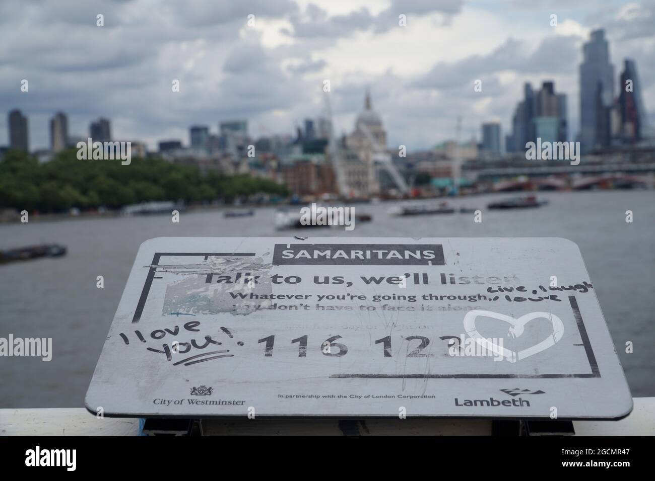 London, Großbritannien, 9. August 2021: Auf der Waterloo Bridge wurde ein Schild, das jeden mit selbstmörderischen Gedanken dazu auffordert, die Hilfslinie der Samariter zu rufen, mit unterstützenden Botschaften der Liebe aufgeschmissen. Die Themse fließt unter der Brücke und die Wolkenkratzer der City of London stehen am Horizont unter einem wolkenlosen Himmel. Anna Watson/Alamy Stockfoto