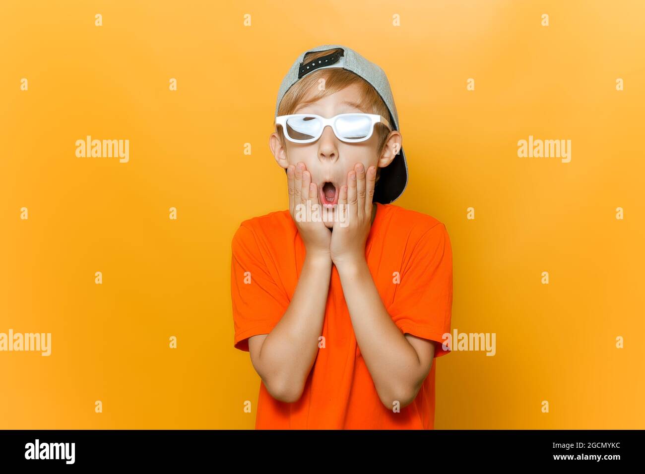 Ein Kind in einer Brille zum Ansehen von Filmen öffnete überraschend seinen Mund und hält seine Wangen mit den Händen Stockfoto