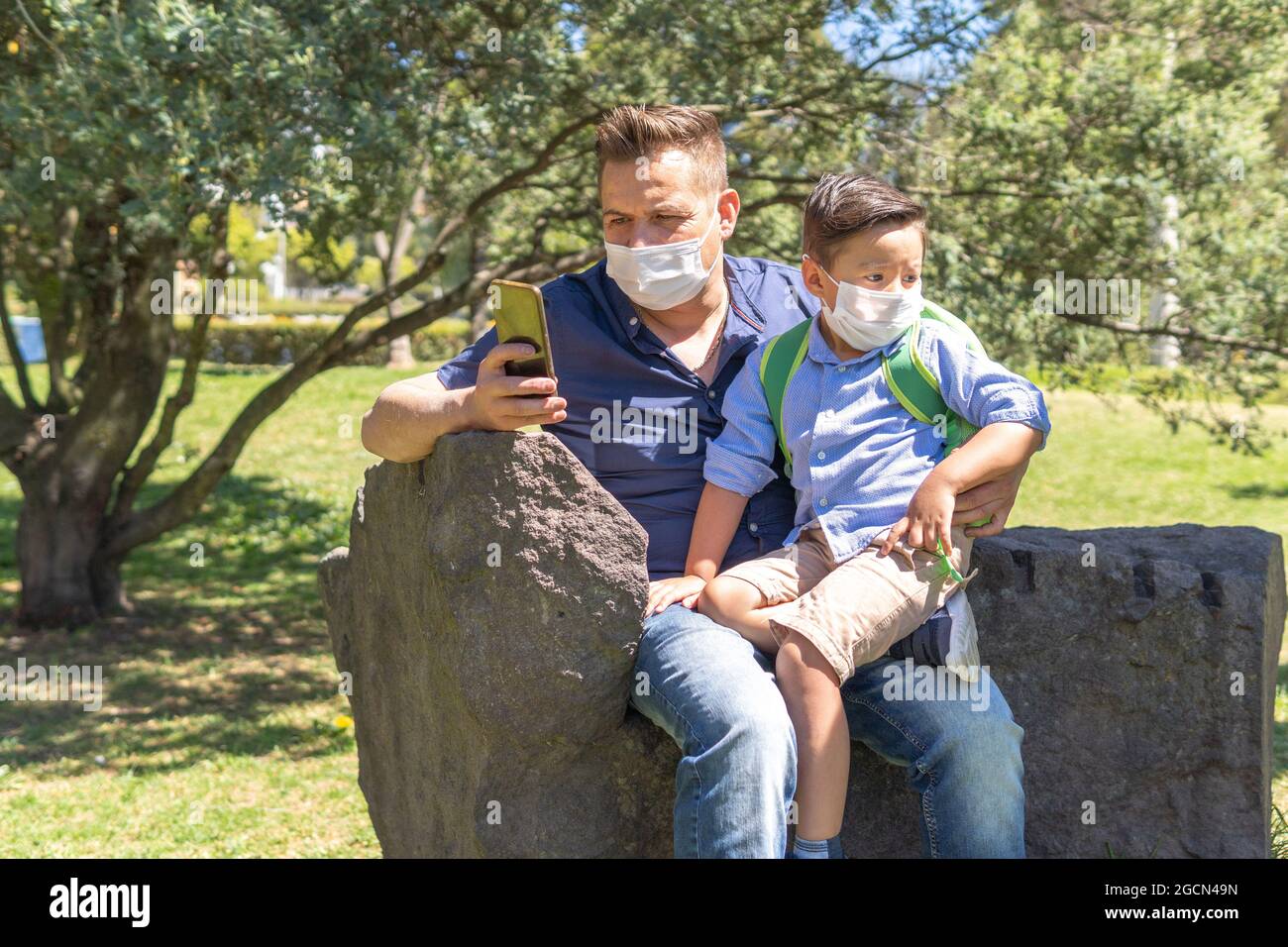 Ein Vater mit einer Maske überprüft sein Handy mit seinem Sohn in den Armen, während sie sich in einem Park ausruhen Stockfoto