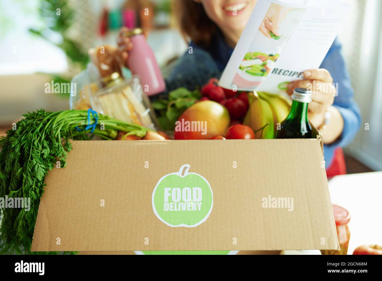 Lieferung von Lebensmitteln. Nahaufnahme einer lächelnden Frau mit einer Lebensmittelbox und einer Rezepturliste in der Küche. Stockfoto