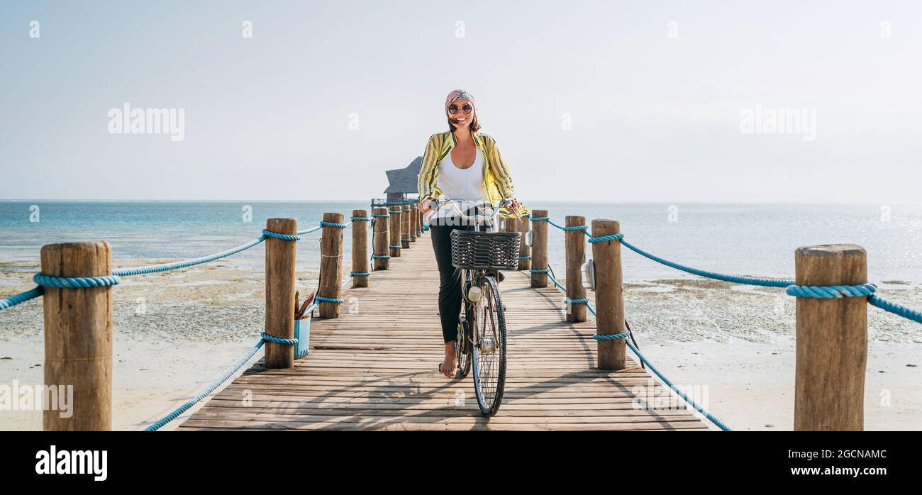 Porträt einer glücklichen lächelnden Frau, gekleidet in leichte Sommerkleidung und Sonnenbrille, die auf dem hölzernen Seebrücke mit dem Fahrrad fährt und die Kamera anschaut. Carele Stockfoto