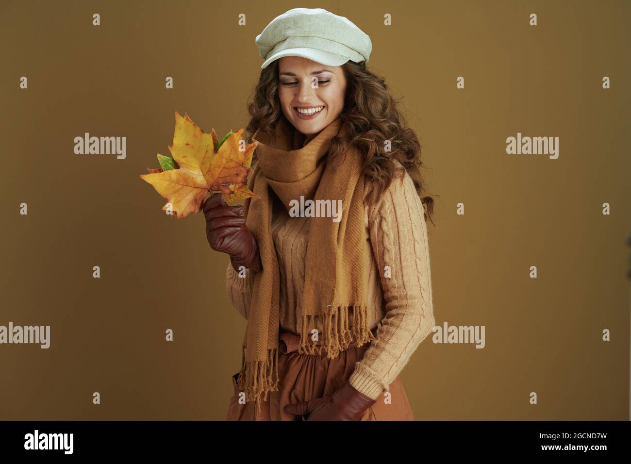 Hallo oktober. Lächelnde Frau mittleren Alters in Pullover mit Lederhandschuhen und gelben Herbst Ahornblätter isoliert auf braunem Hintergrund. Stockfoto