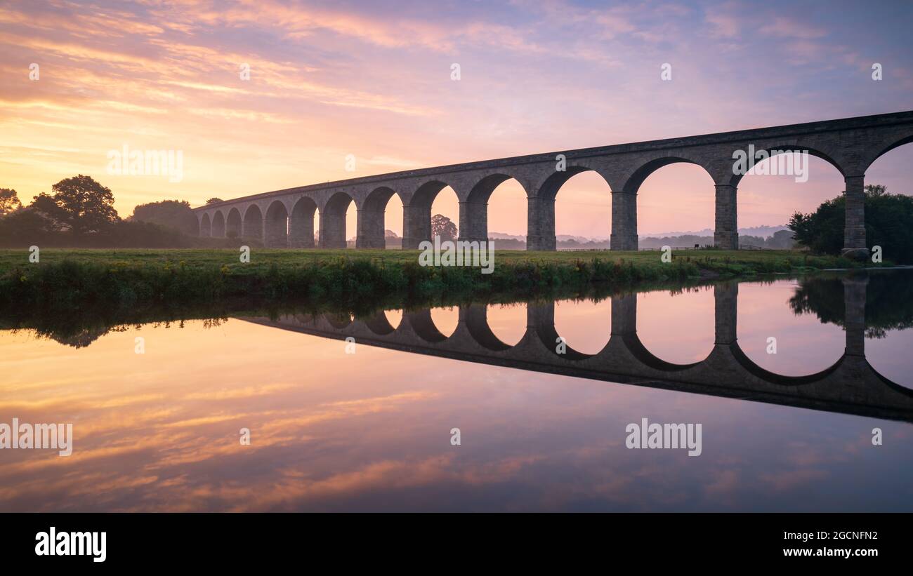 Ein schöner Sommersonnenaufgang umrahmt die Bögen des Arthington Viadukts an einem ruhigen und warmen Augustmorgen, wobei sich die Szene im River Wharfe widerspiegelt. Stockfoto