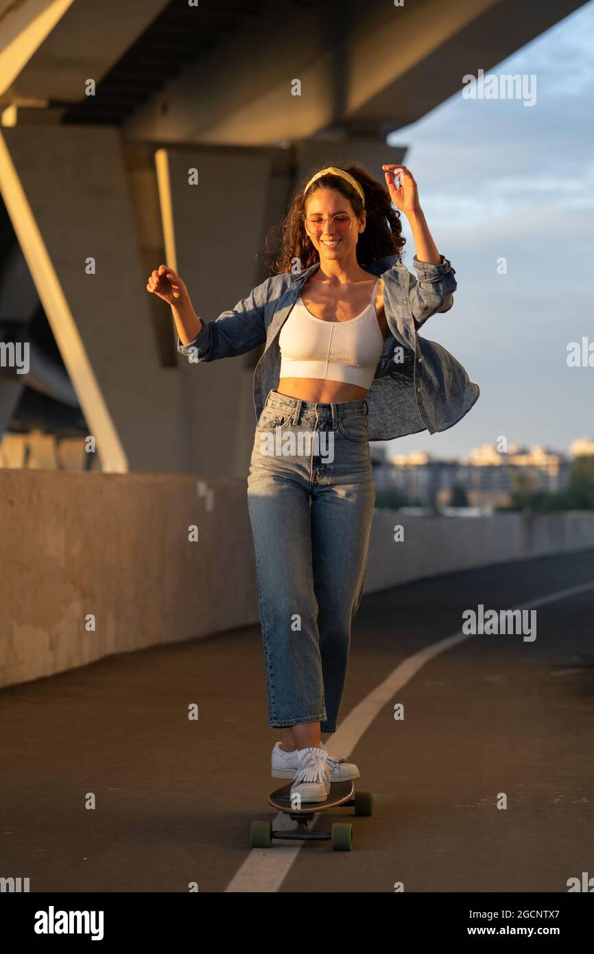 Junge unbeschwerte Frau genießt es, bei Sonnenuntergang Skateboard zu fahren. Glückliches Mädchen in trendigen Street-Style-Kleidung Stockfoto