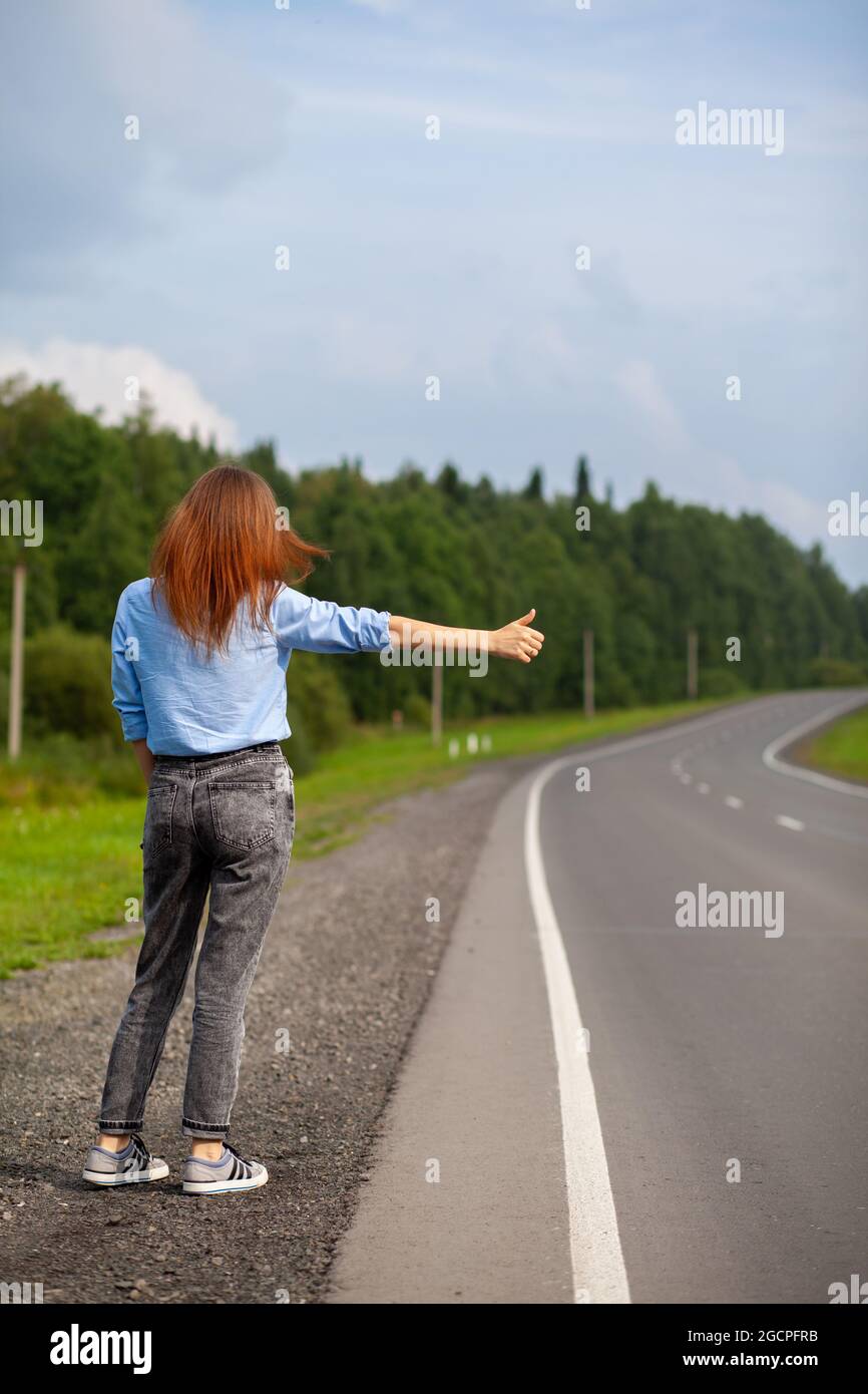 Das Mädchen hält das Auto mit der Hand auf der Autobahn an. Stilvolle Frau auf der Straße hält das Auto auf eine Reise gehen. Eine Straße mitten im Wald. Stockfoto