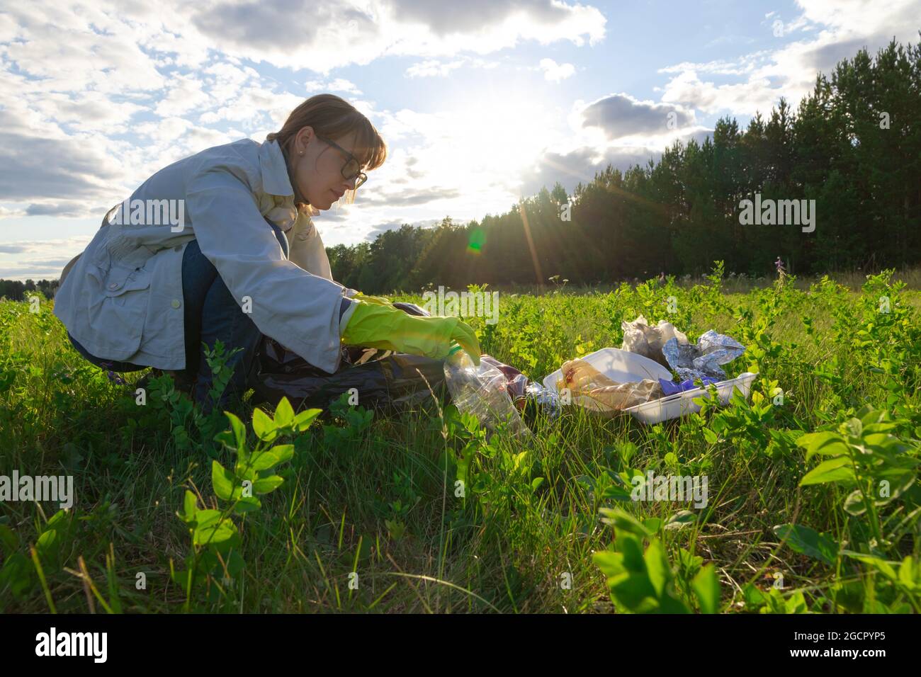 Junge Frau in Gummihandschuhen auf der Natur sammelt Plastikmüll an einem sonnigen Sommertag vor dem Hintergrund von Bäumen. Umweltschutz. Auswahl Stockfoto