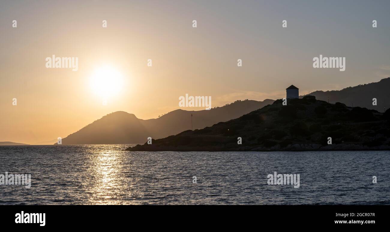 Alte Windmühle auf einem Felsen mit griechischer Flagge, Sonnenuntergang, Panormitis Bay, Simi, Dodekanes, Griechenland Stockfoto