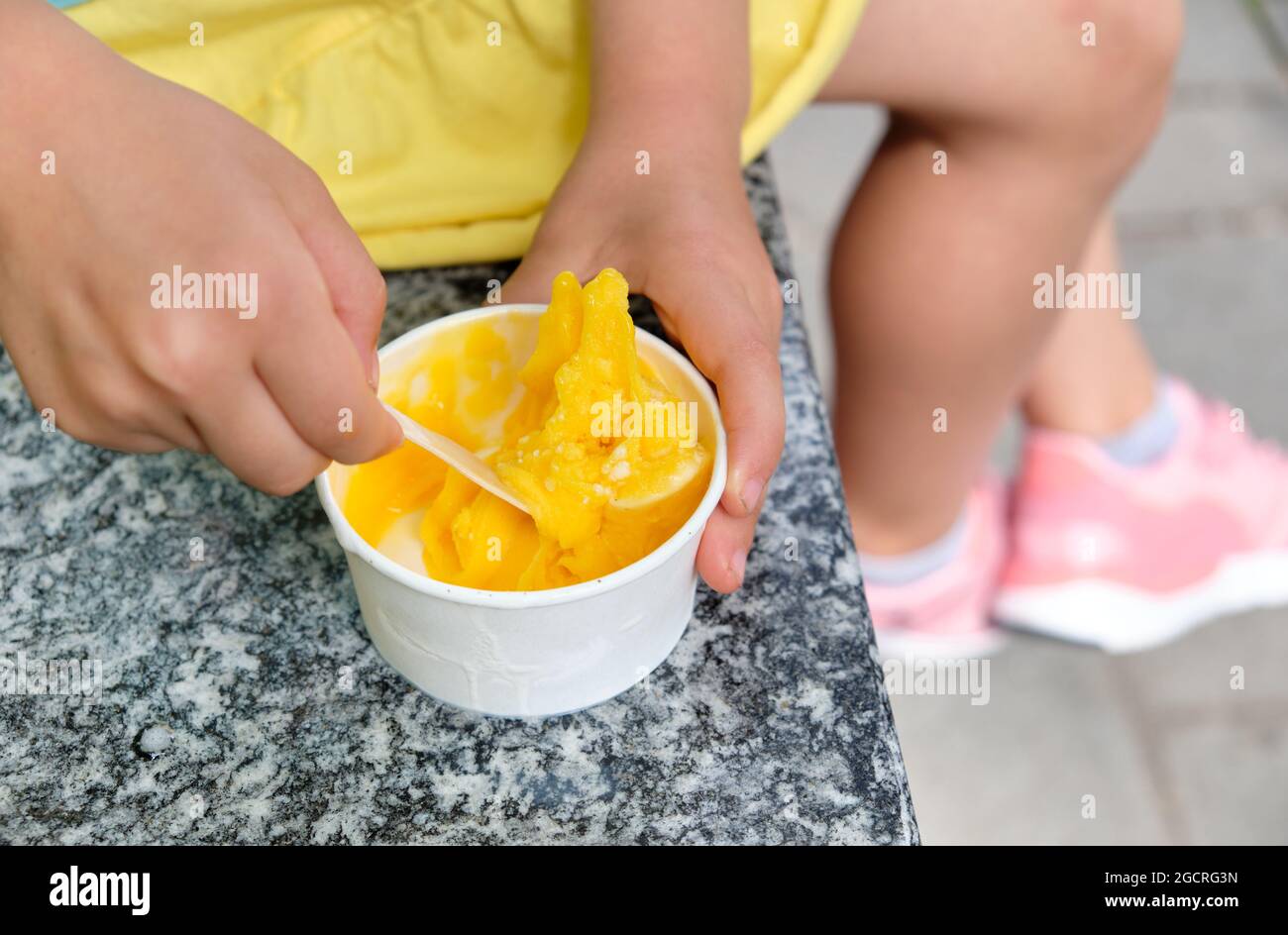 Nahaufnahme eines 5-jährigen kaukasischen Mädchens in Sommerkleidung, das sitzt und aus einer Tasse gelbes Mango-Eis isst. Gesehen in Deutschland im Juni Stockfoto