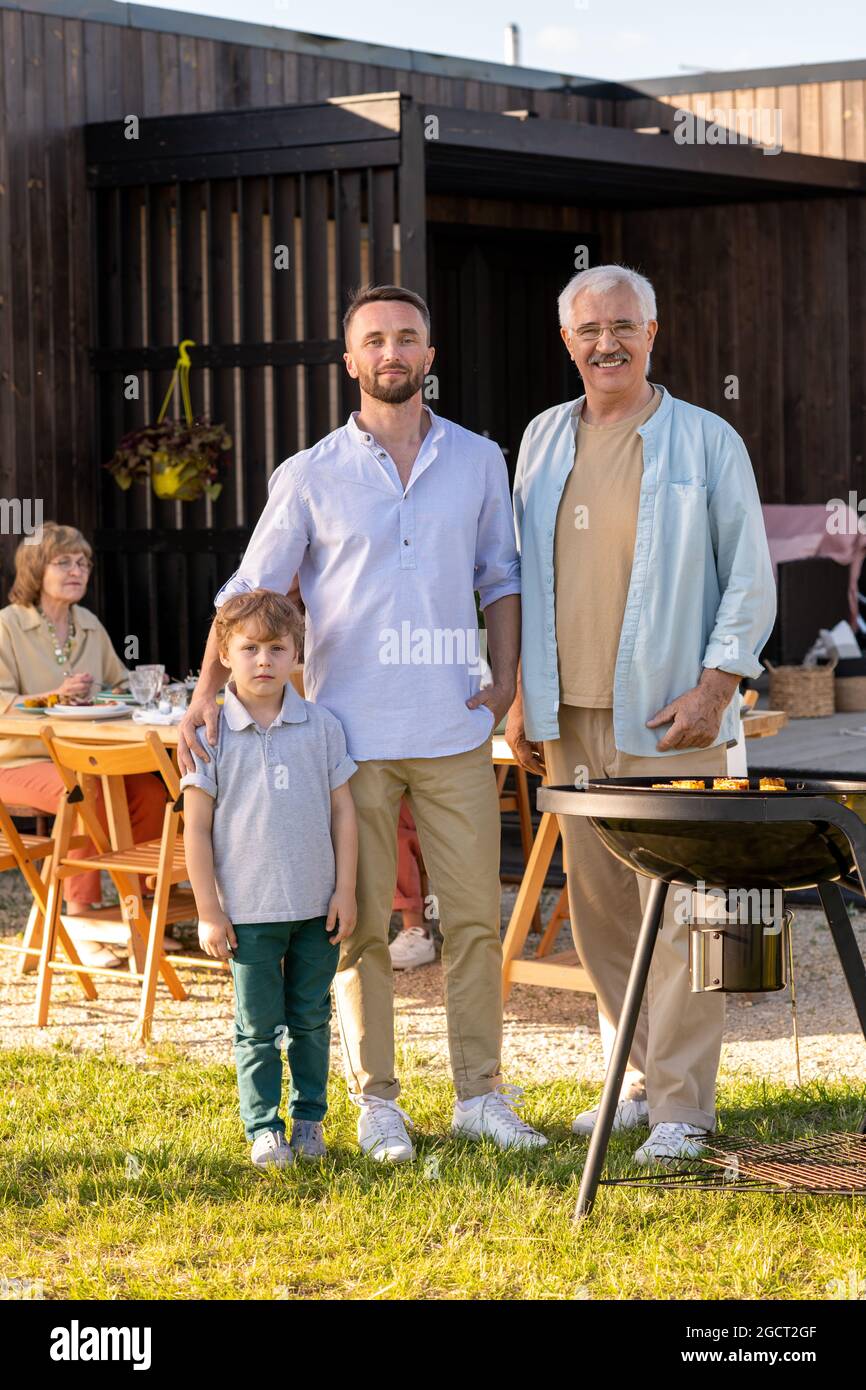 Drei Männer in Hosen und blauen Hemden stehen auf dem grünen Rasen beim Grill während des Familien-Picknicks Stockfoto
