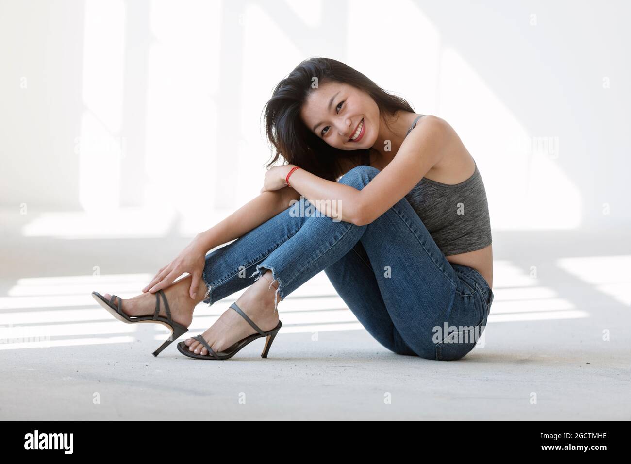 Portrait schöne asiatische Mädchen auf dem Boden sitzen. Sie trägt blaue Jeans, ein sportliches Top und Stiletto Heels. Dahinter kommt das Sonnenlicht. Stockfoto