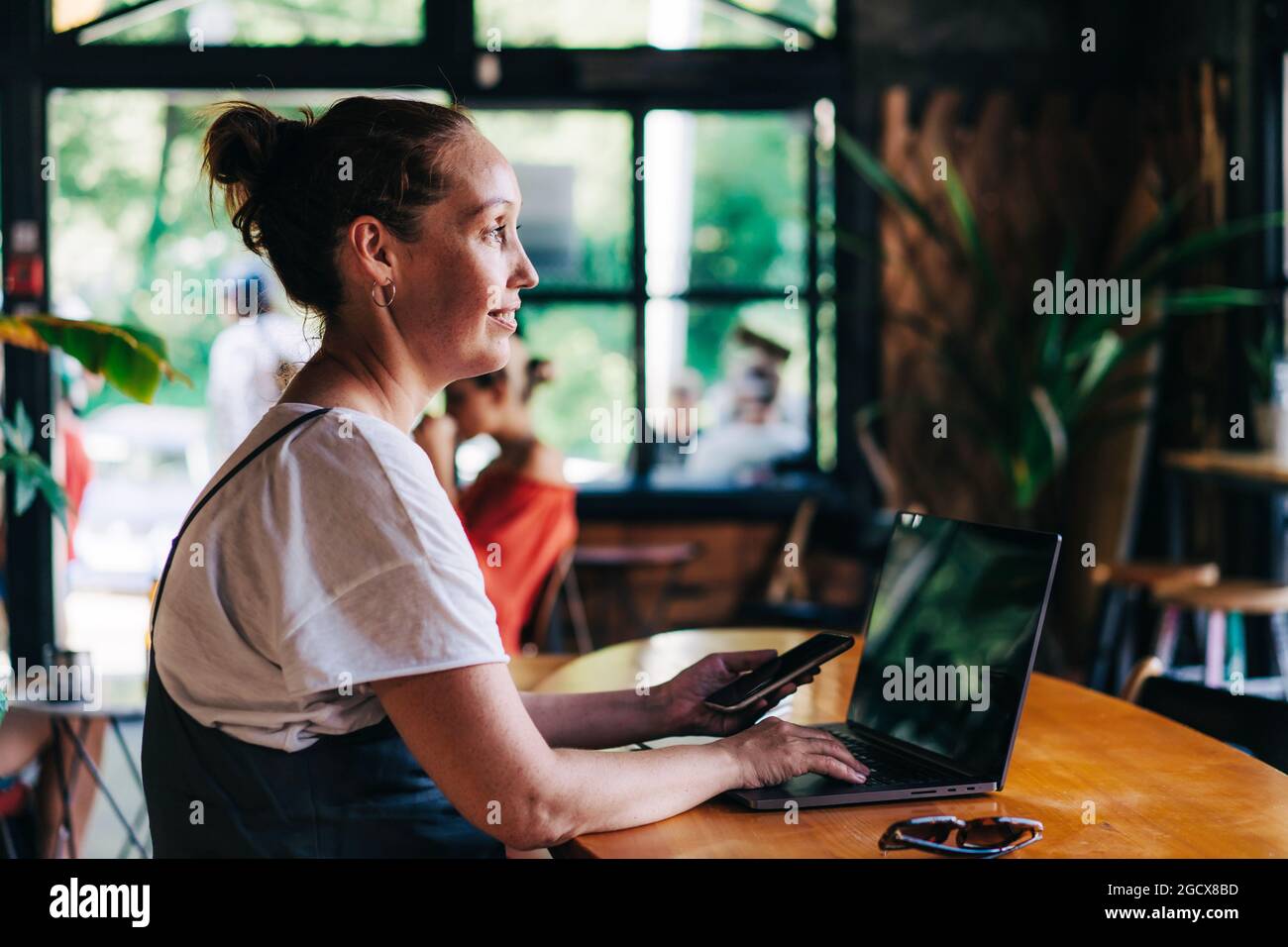 Eine niedliche, inspirierte Frau, die mit drahtlosen Technologien von einem gemütlichen Café aus ferngesteuert auf einem Laptop arbeitet. Stockfoto