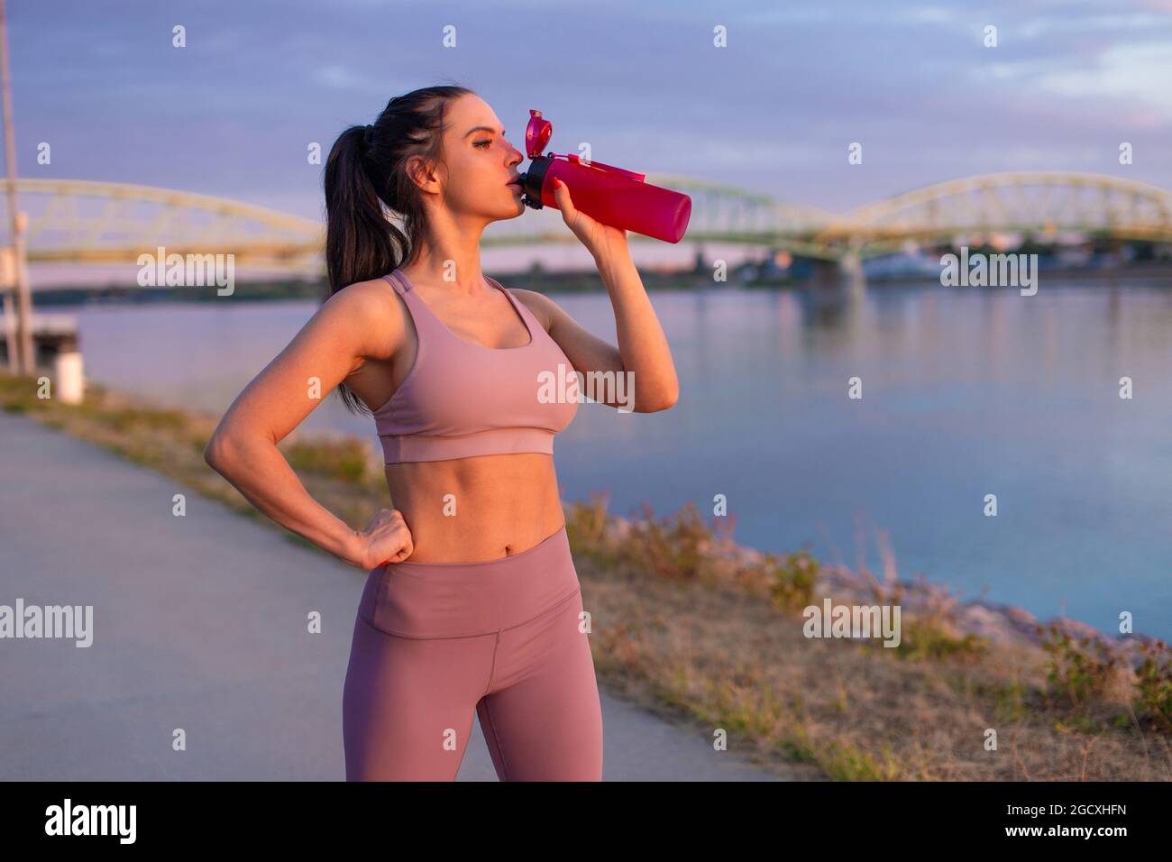 Durstige junge Frau in Sportkleidung, die am Flussufer Erfrischung aus der Flasche trinkt Stockfoto