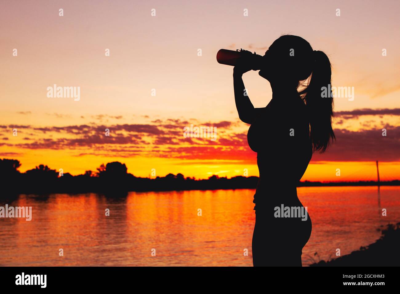 Durstige junge Frau Silhouette trinken Erfrischung aus der Flasche am Flussufer in Sonnenuntergang Stockfoto