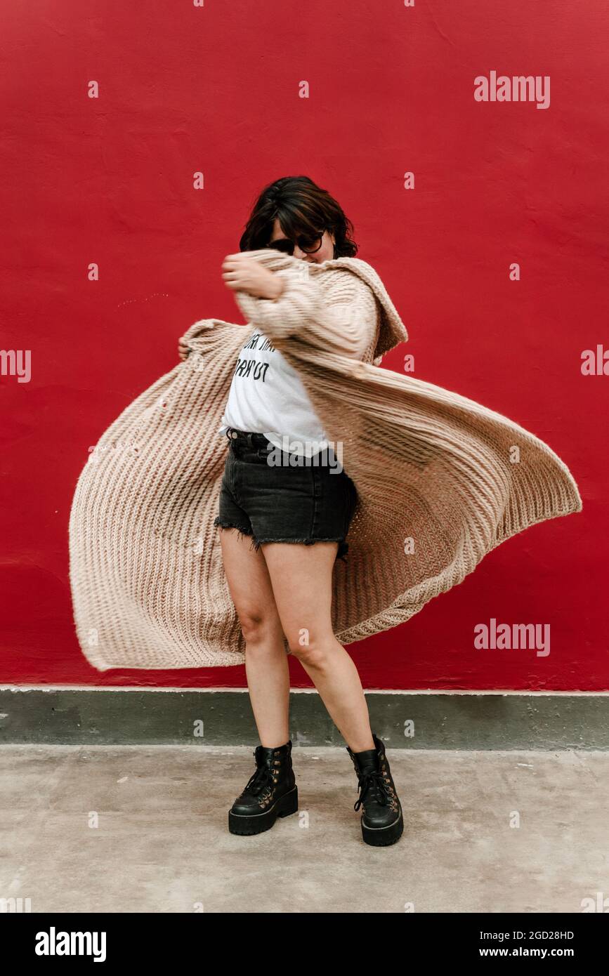 Bild einer Frau, die mit ihrem Pullover spielt und es fliegen lässt. Selektive Fokussierung der Frau beim Drehen mit ihrem handgestrickten Pullover auf rotem Hintergrund Stockfoto