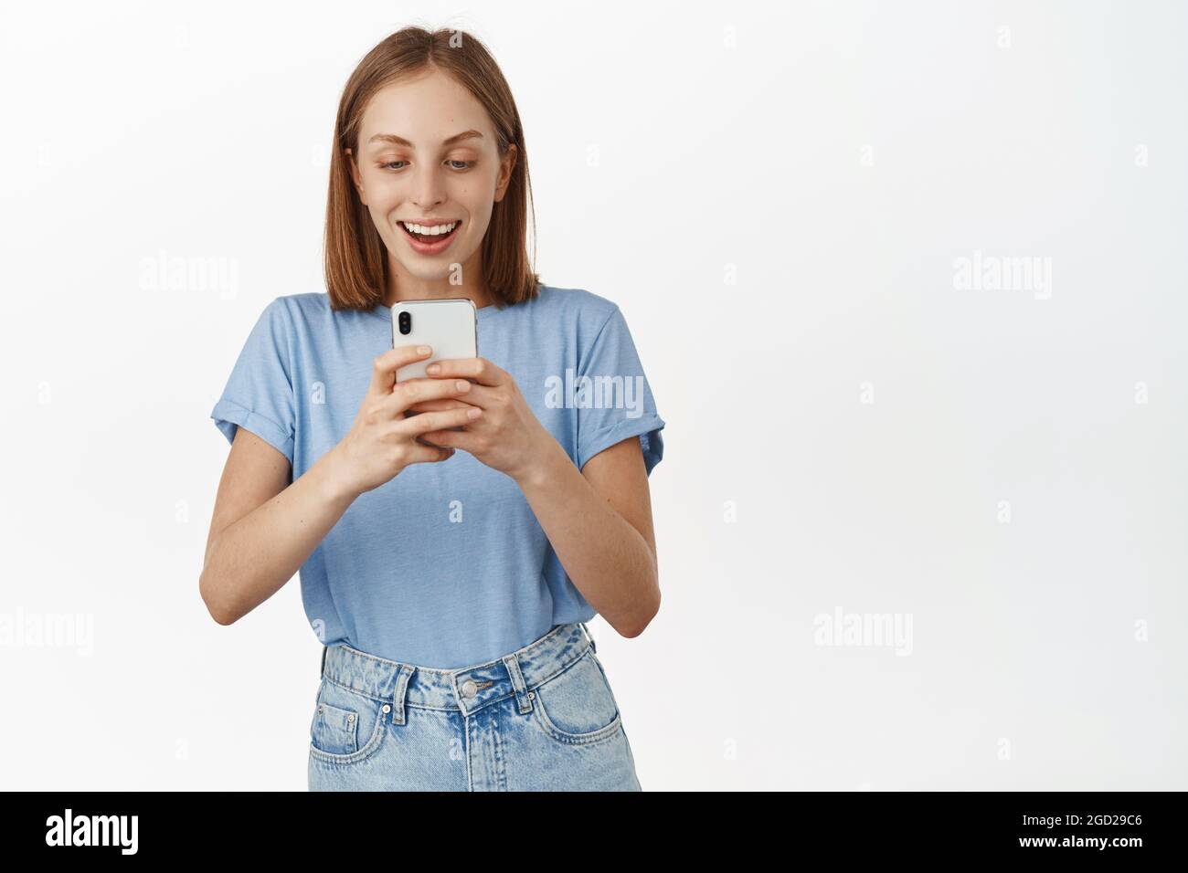 Aufgeregt lächelndes blondes Mädchen, das Foto auf dem Handy. Junge glückliche Frau, die auf dem Smartphone-Bildschirm staunt, fand smth im Online-Shop, stehend Stockfoto