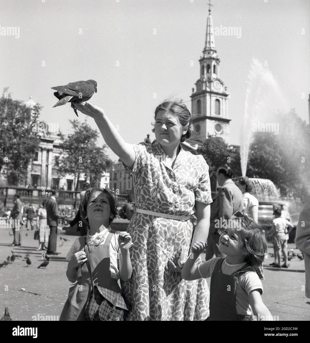 1950, historisch, tagsüber und eine Mutter mit ihren zwei kleinen Kindern am Trafalgar Square, London, England, Großbritannien. Die Dame hat eine Taube auf der Handfläche ihrer ausgestreckten Hand sitzend, während ihre beiden jungen Töchter vor Ehrfurcht ihre tapfere Mutter betrachten. Stockfoto