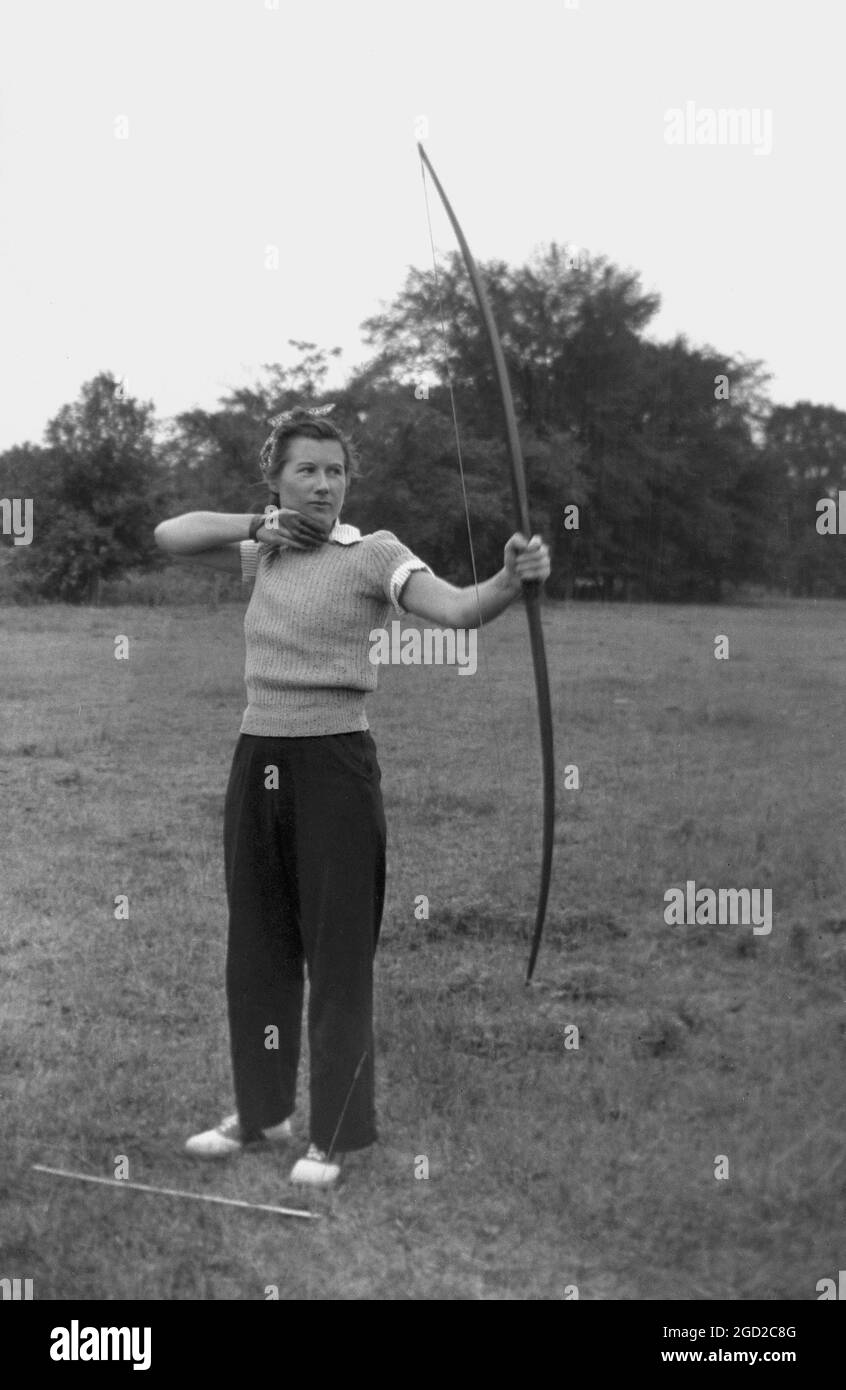 1950er Jahre, historisch, draußen auf einem Feld, eine Frau, die ein Paar Slacks trägt, die mit Pfeil und Bogen stehen und Bogenschießen machen. Sowohl eine sportliche als auch eine lustige Freizeitbeschäftigung, im Bogenschießen benutzt man einen Bogen, um zu versuchen und Pfeile so nah wie möglich an der Mitte eines Ziels zu schießen. Stockfoto