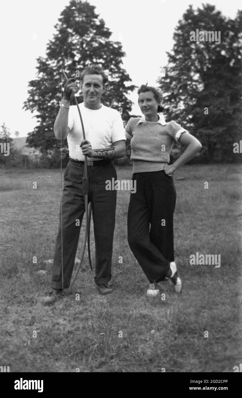 1950er Jahre, historisch, draußen auf einem Feld, eine Bogenschützin und ihr männlicher Lehrer. Das Bogenschießen ist sowohl eine Sport- als auch eine Freizeitbeschäftigung, um Pfeile so nah wie möglich an der Mitte eines Ziels zu schießen. Stockfoto