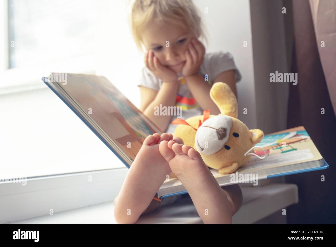 Niedliches kleines kaukasisches Kind sitzt neben dem Fenster und liest Buch. Stockfoto