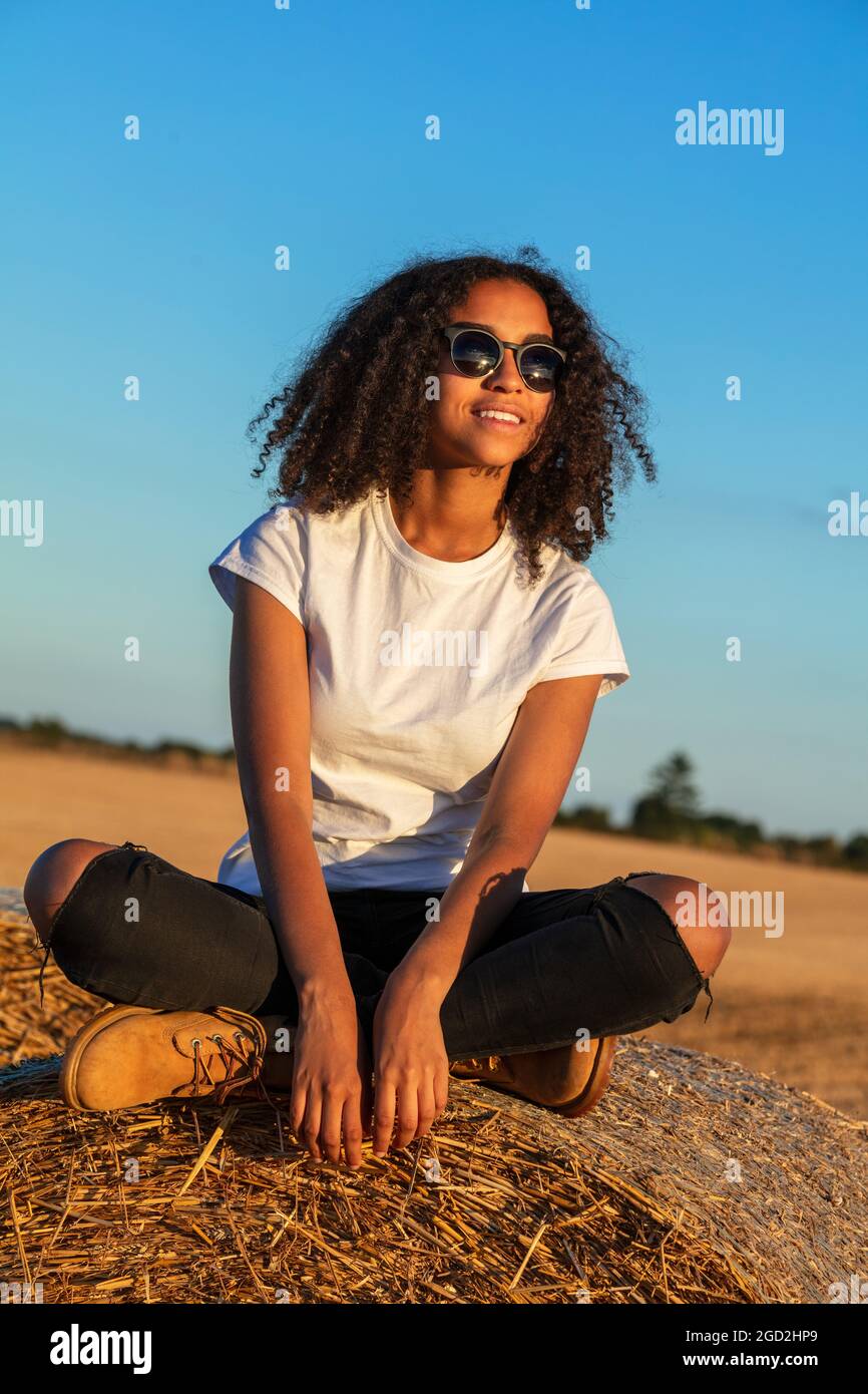 Schöne gemischte Rasse afroamerikanische weibliche Teenager junge Frau sitzt auf einem Heuballen in einem Feld in weißen T-Shirt und Sonnenbrille bei Sonnenuntergang Stockfoto