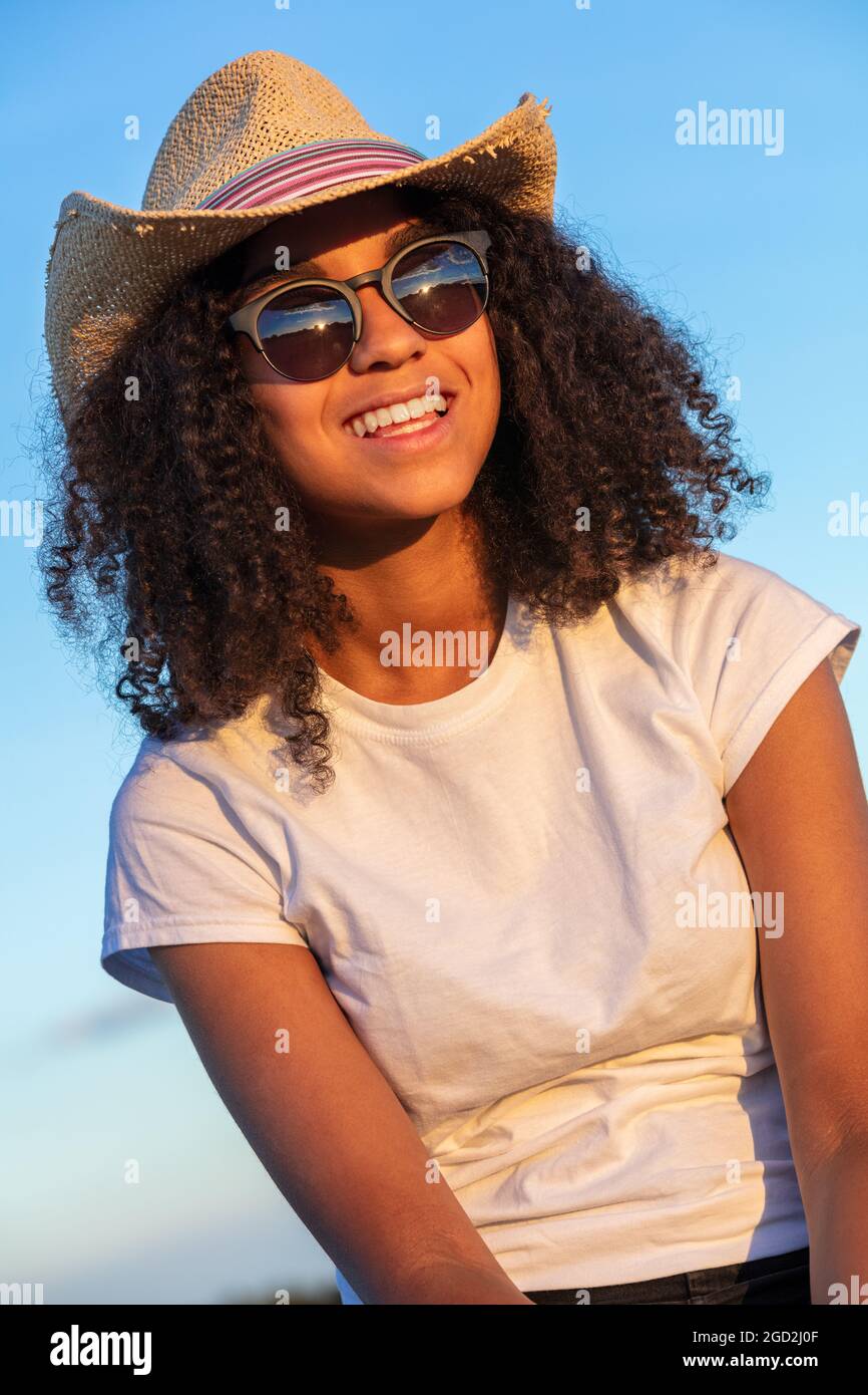 Schöne und glückliche gemischte Rasse afroamerikanische weibliche Mädchen Teenager junge Frau mit perfekten Zähnen tragen Stroh Cowboy Hut Sonnenbrille und weißen t-sh Stockfoto