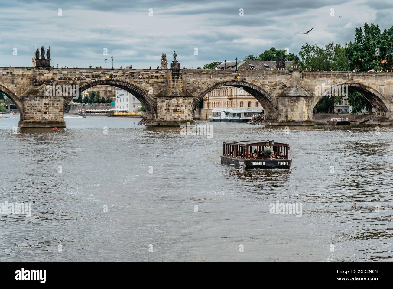 Prag, Tschechische Republik - August 4,2021. Kleine hölzerne Fähre Boot mit Passagieren überqueren Moldau, Karlsbrücke im Hintergrund. Lokalen Wassertransport Stockfoto